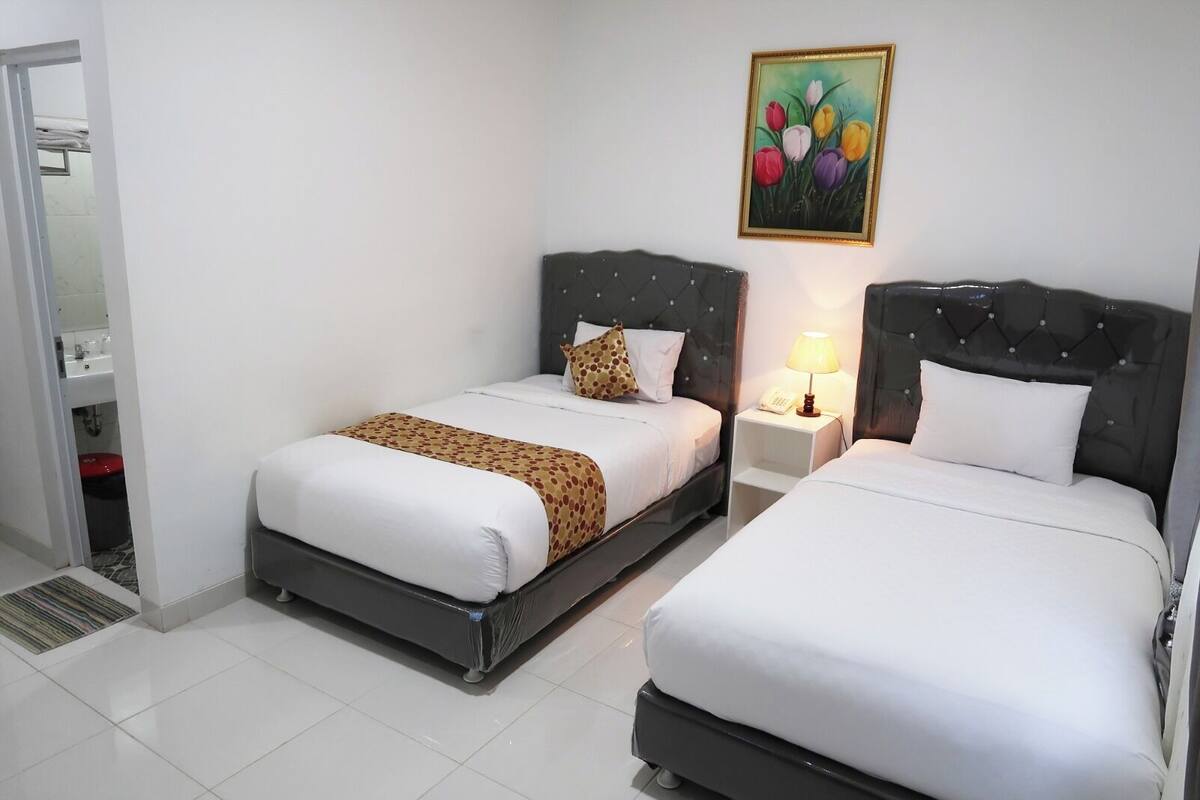 A hotel room with two beds, a painting, a bedside lamp, and a bathroom visible through an open door.