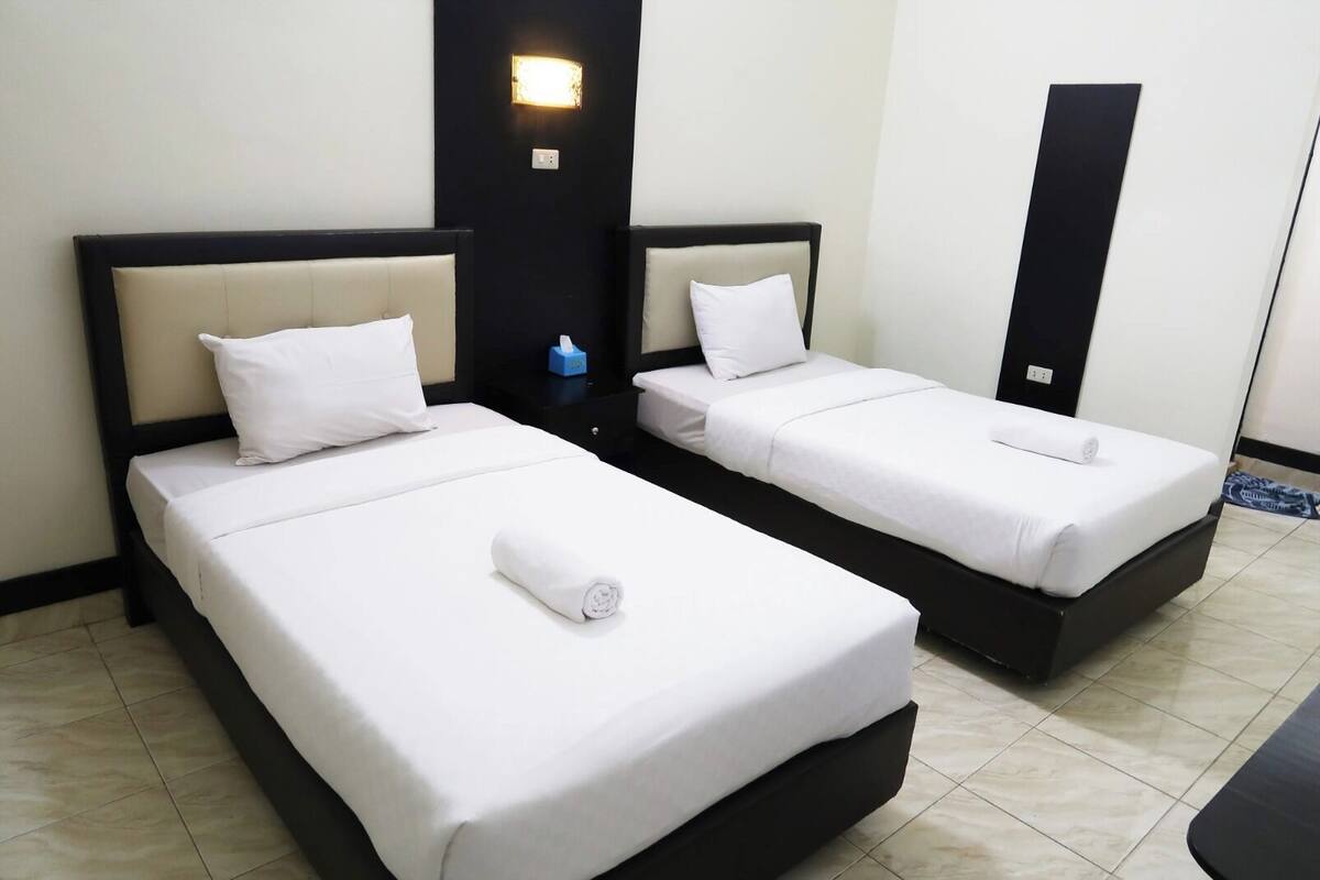 Two single beds with white linens, a nightstand with a lamp, and a black vertical object on the wall.