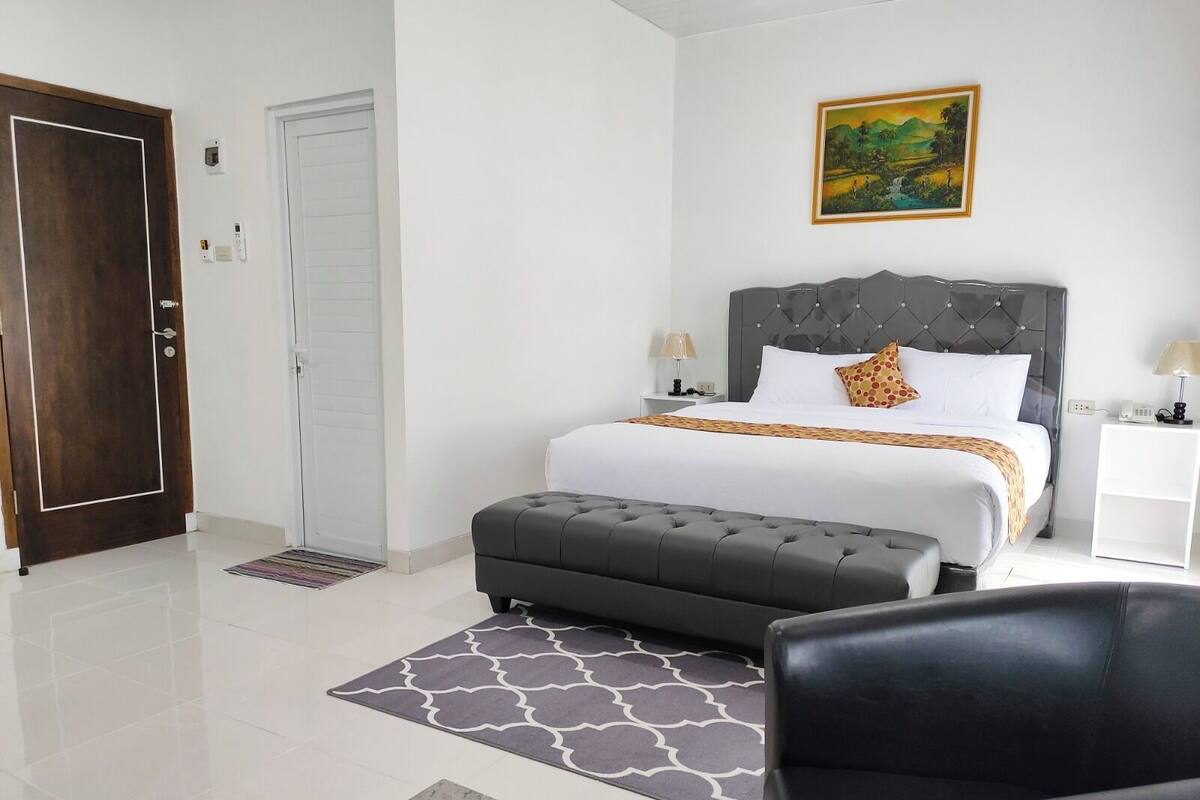 A bedroom with a bed, a grey upholstered bench, a white bedside table with a lamp, a door, and a wall-mounted painting.