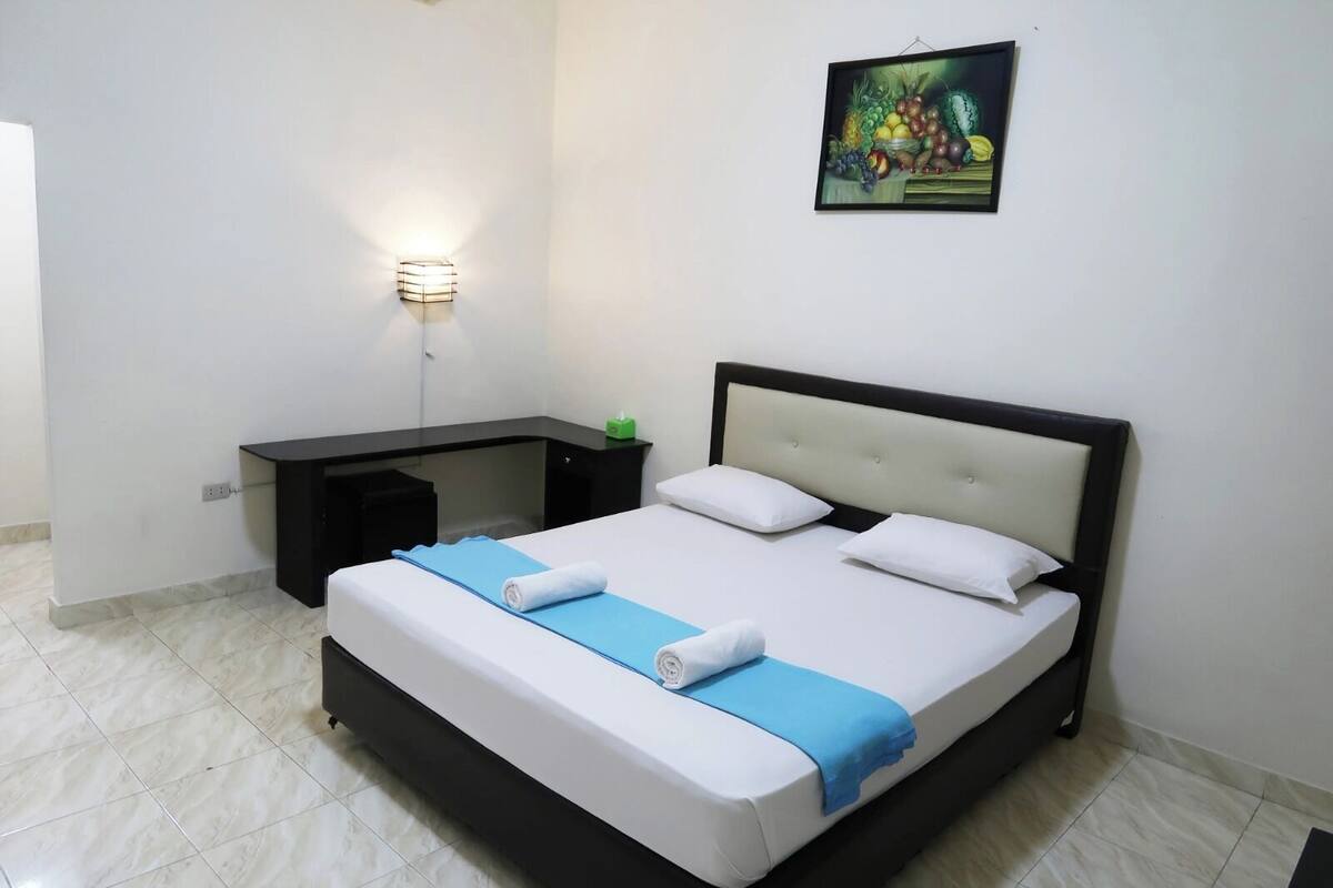 A hotel room with a bed, a nightstand, a wall-mounted light, and a fruit painting.