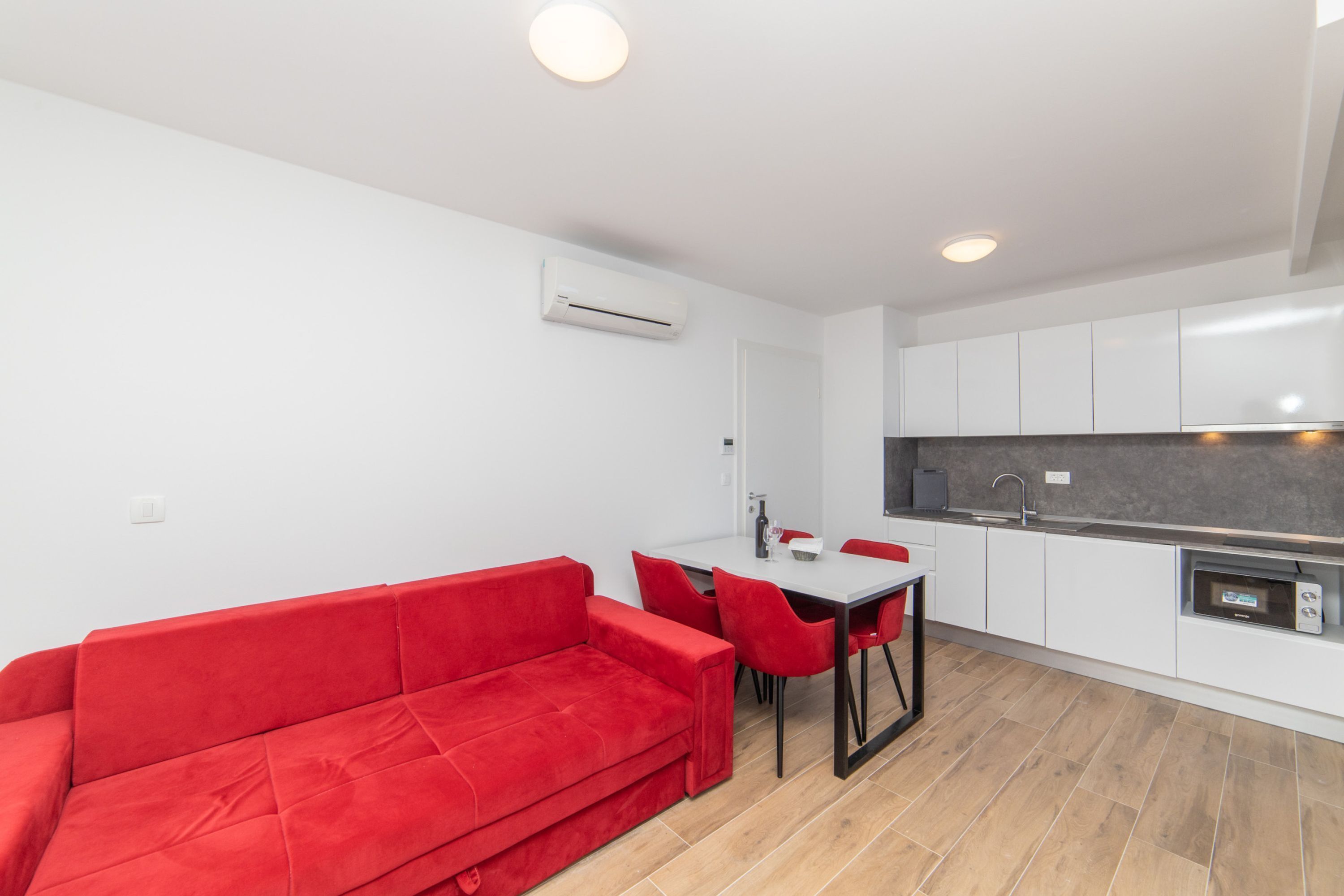 Un espacio habitable pequeño con un sofá rojo, una mesa de comedor con sillas, una kitchenette y muebles de cocina blancos.
