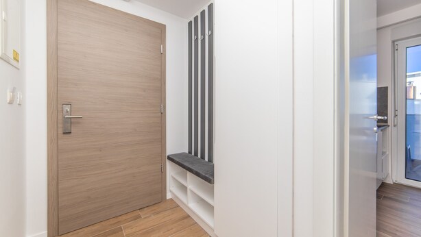 Een gang met een houten deur, een bankje en een witte muur.