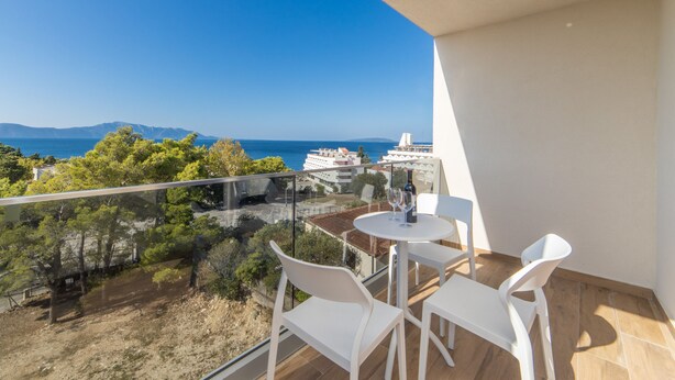 A balcony with a table and chairs, overlooking a coastal area with buildings and trees. - Altiora Prestige Residence 101 (Gradac)
