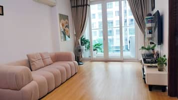 A living room with a sofa, a standing fan, a potted plant, and a window with curtains.