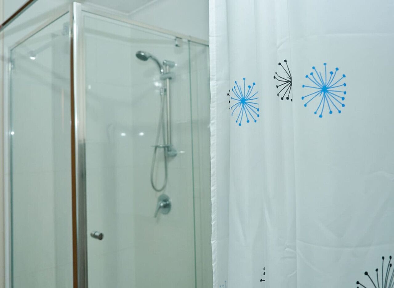 A glass-enclosed shower with a handheld showerhead, a glass door, and a white curtain with abstract blue and black designs.