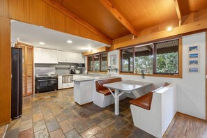 A modern kitchen with white cabinetry, a central island, and a dining area with a table and benches. - Multi-Family Coastal Escape | Camp & Stay (St Leonards)