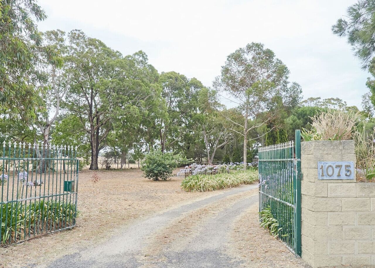 A gated property entrance with a dirt path leading to a building, surrounded by trees and a sign with the number 1075.