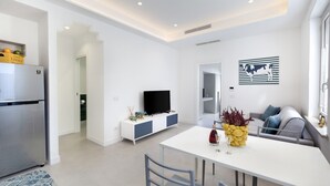 A modern kitchen with a built-in refrigerator, a dining area with a white table and chairs, a gray sofa, and a flat-screen TV on a white cabinet. - CASA APREDA (Sorrento)
