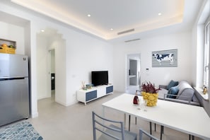 A modern kitchen with a built-in refrigerator, a dining area with a white table and chairs, a gray sofa, and a flat-screen TV on a white cabinet. - CASA APREDA (Sorrento)