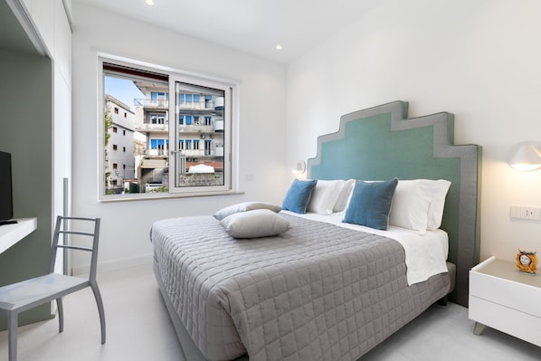 A bedroom with a bed, a chair, a nightstand, a lamp, and a window with a view of buildings. - CASA APREDA (Sorrento)