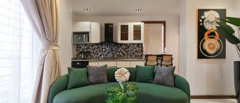 A living room with a green sofa, a coffee table, and a plant. There is a kitchen in the background with cabinets and a tiled backsplash.