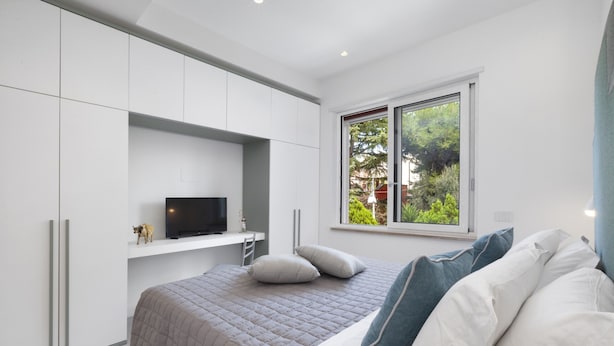 A bedroom with a bed, a TV on a shelf, a window with a view of greenery, and white cabinetry. - Stayxperience - Casa Apreda (Sorrento)