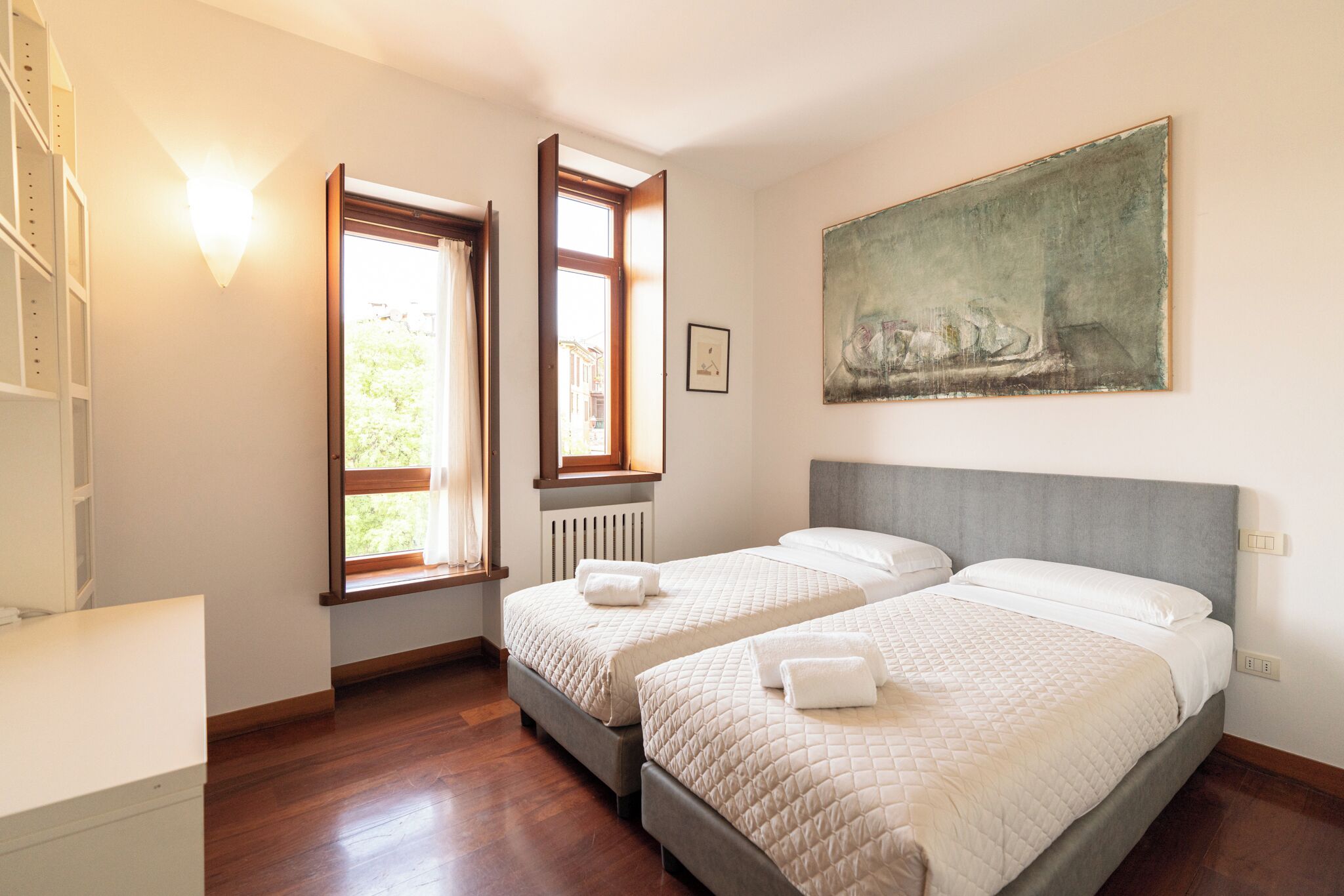A hotel room with two beds, a painting on the wall, and a window with curtains.