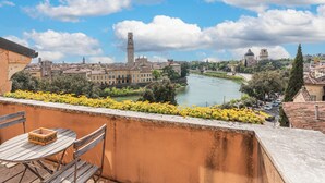 A balcony with a table and chairs overlooking a river, historic buildings, and a tower. - Residenza vista mozzafiato Ponte Pietra (Verona)