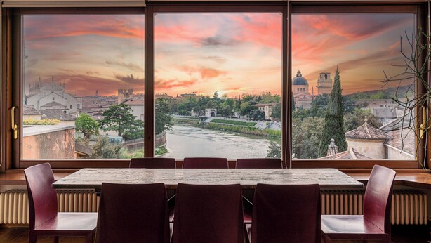 A dining area with a table and chairs, a view of a river, and buildings in the background. - Residenza vista mozzafiato Ponte Pietra (Verona)