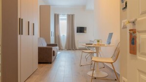 A modern hotel room with a sofa, a dining table with chairs, a TV, and a small plant.