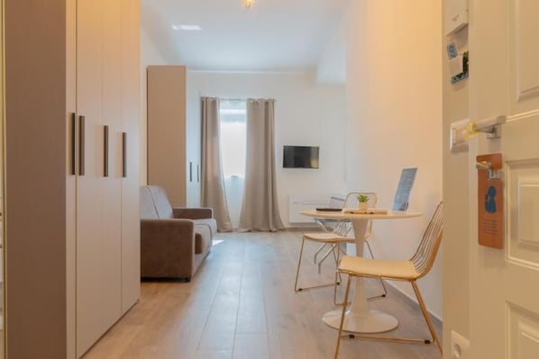 A modern hotel room with a sofa, a dining table with chairs, a TV, and a small plant.