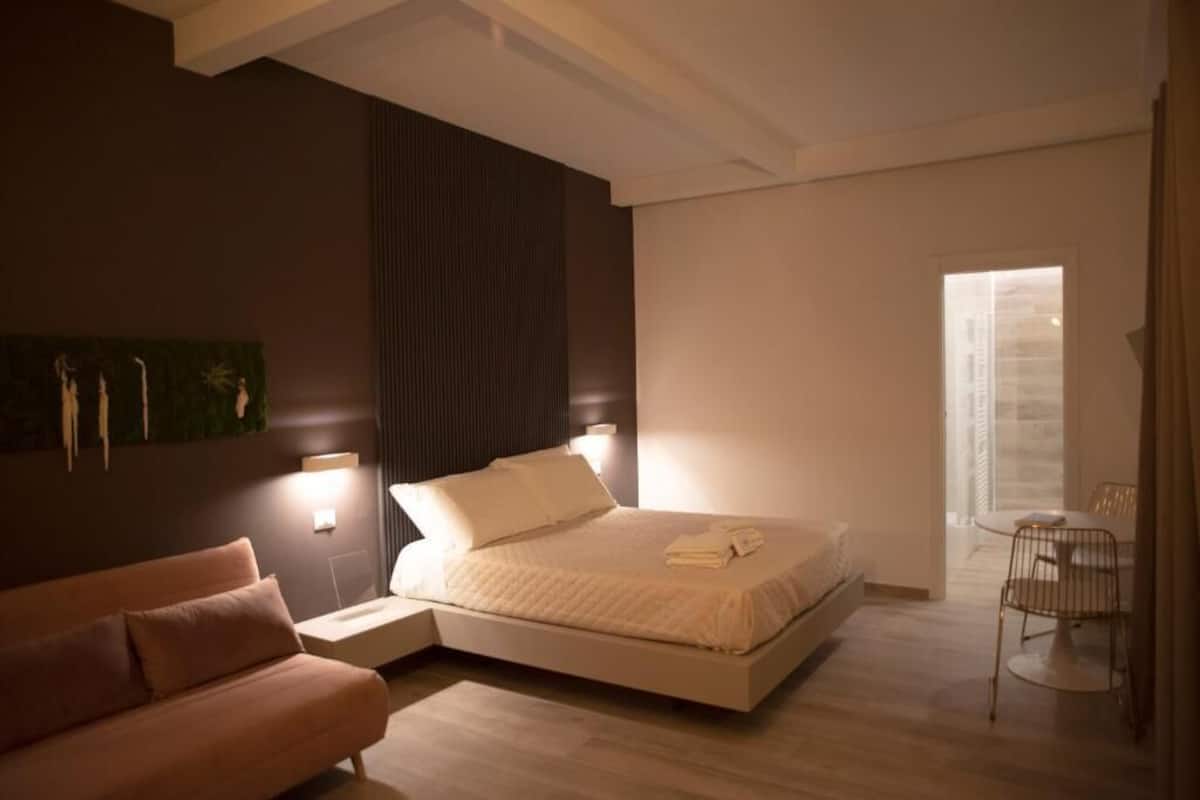 A modern bedroom with a bed, bedside tables, a sofa, and a dining area.