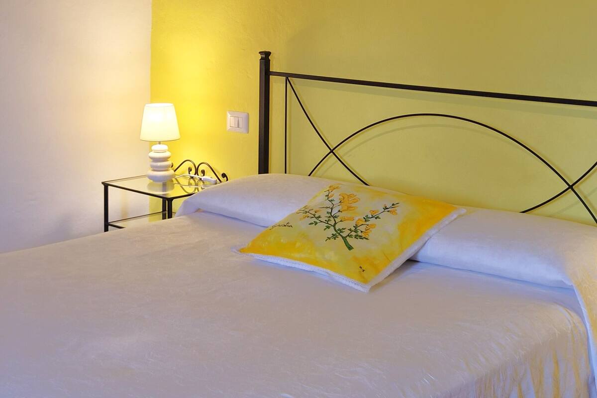 Una cama con colcha blanca y almohada floral amarilla, una mesita de noche con lámpara, y una pared con detalle en color amarillo.
