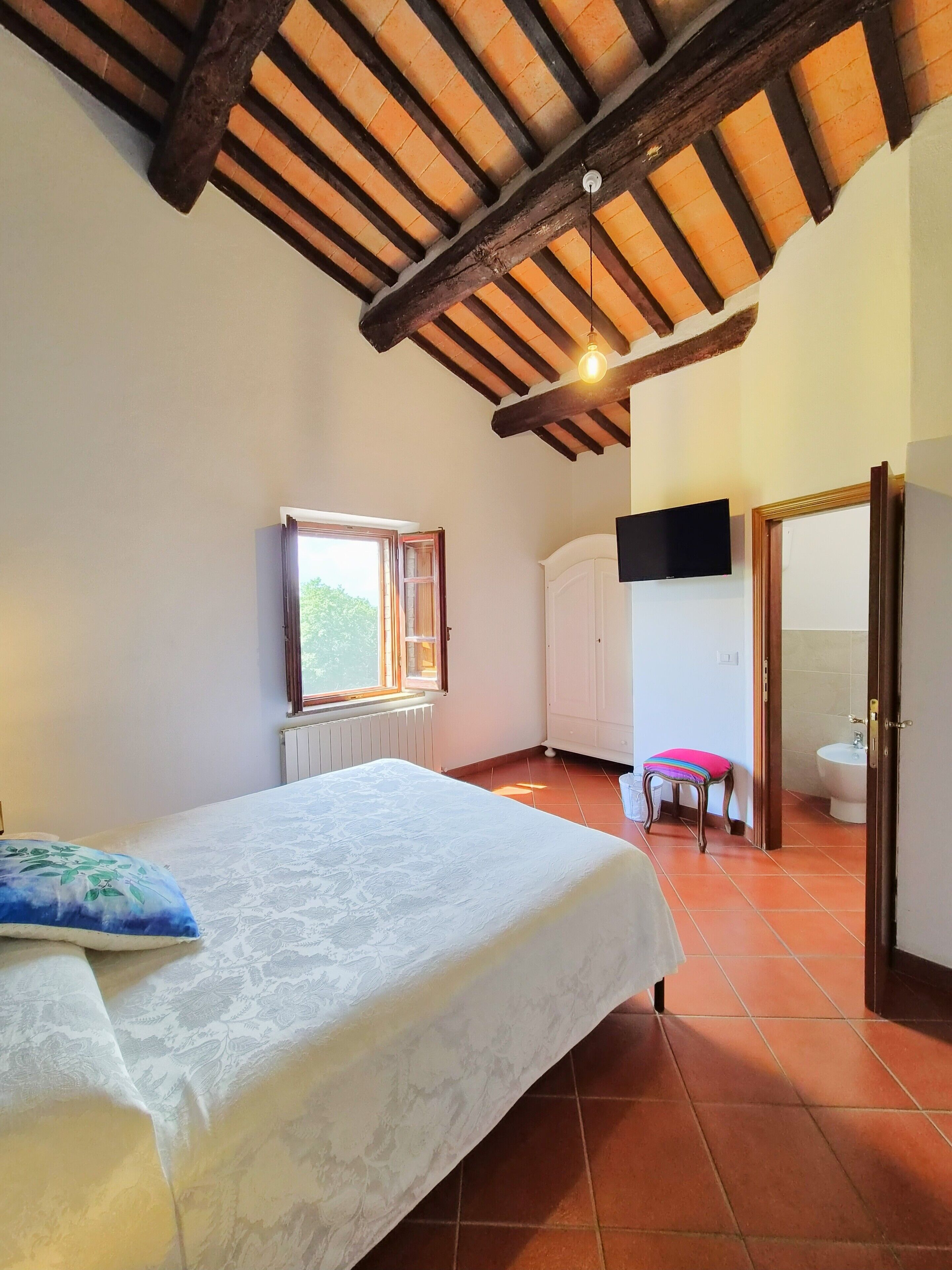 Un dormitorio con cama, ventana, televisor y baño visible a través de una puerta abierta.