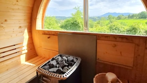 A wooden sauna with a large arched window, a charcoal bin, and a wooden bucket.
