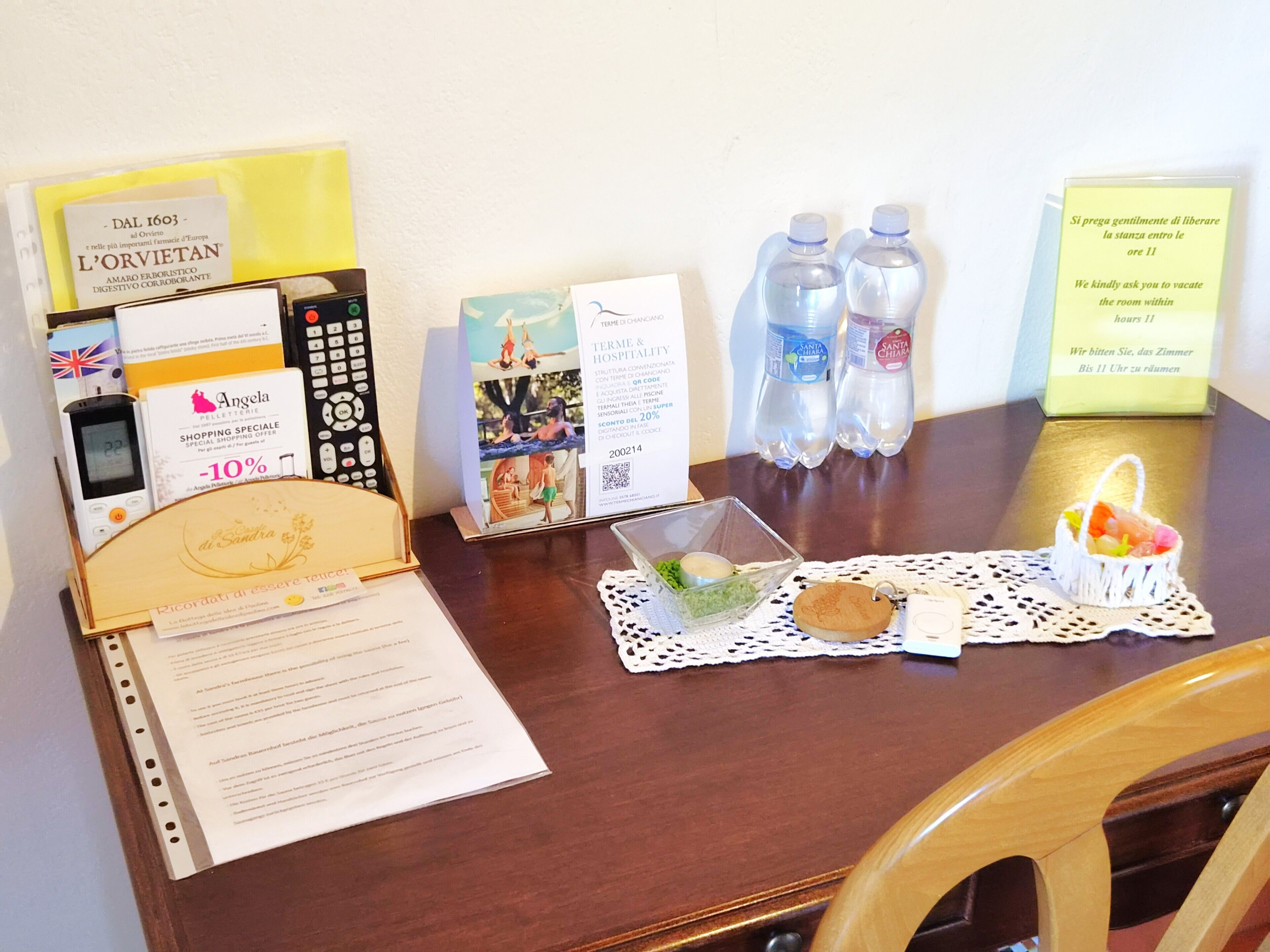 A hotel room desk with a remote control, a mobile phone, a water bottle, a tray with a candle, and informational cards.
