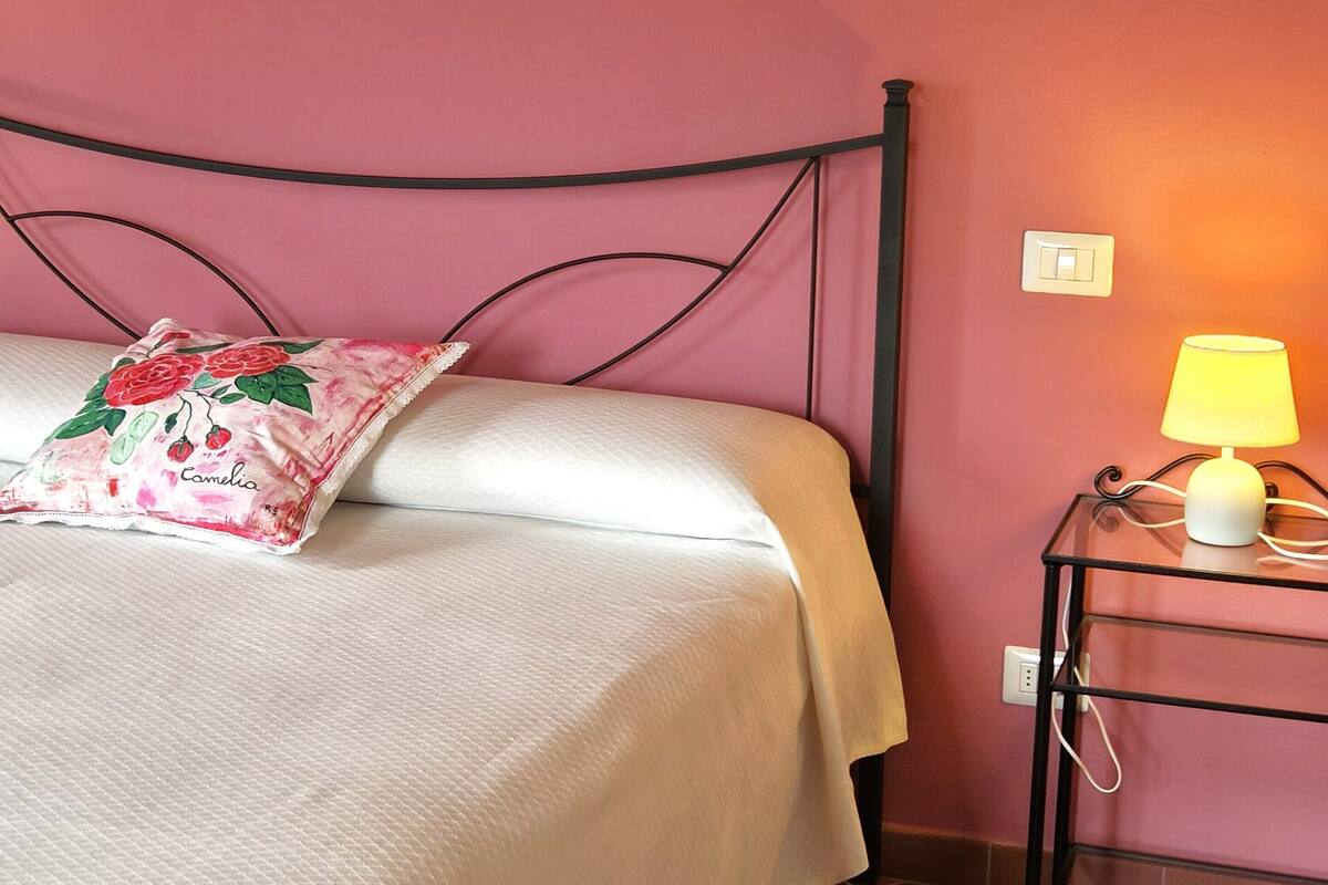 Una cama con cubrecama blanco y almohada floral, una mesita de noche con lámpara, e interruptor de pared.