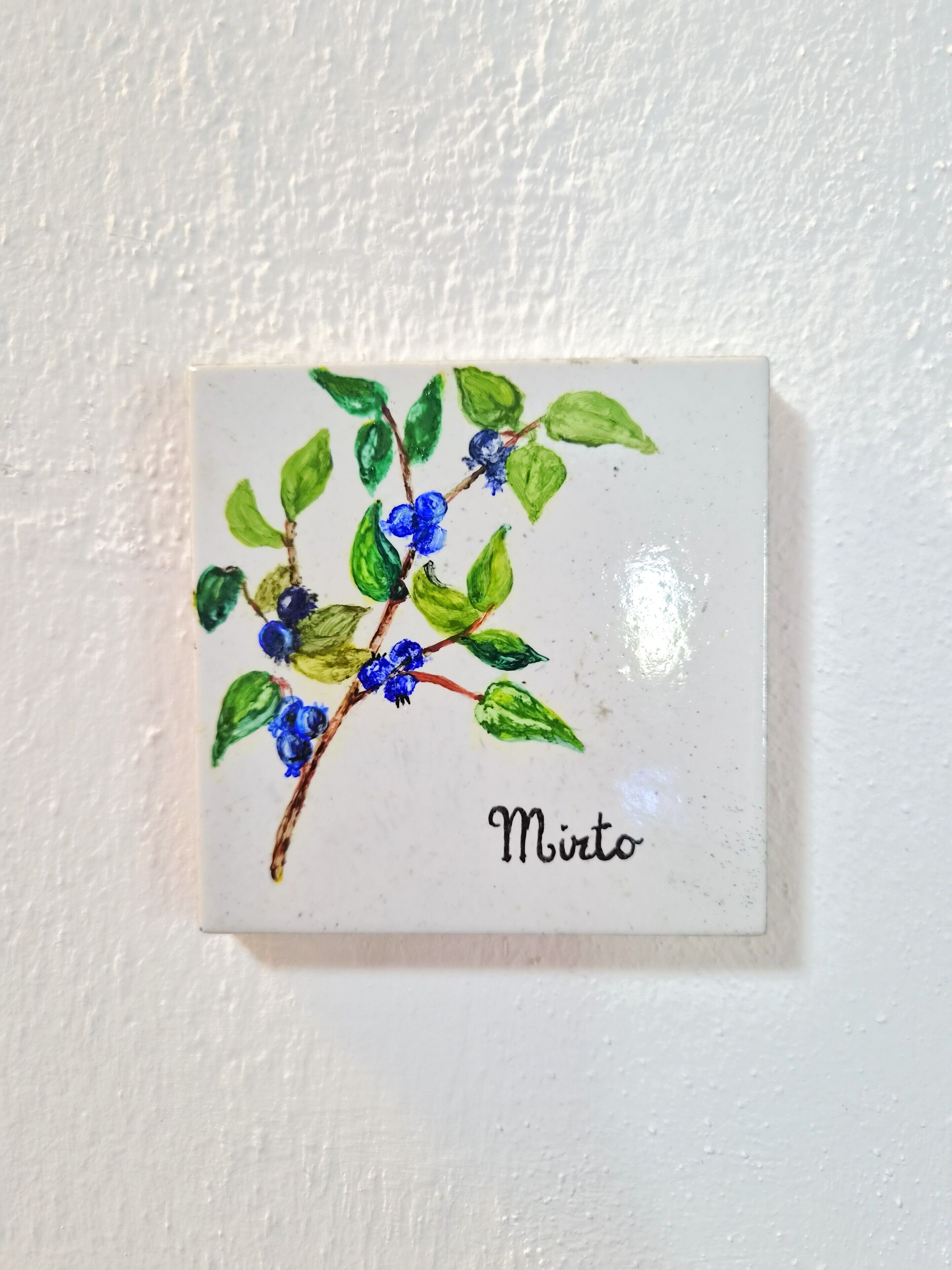 A small, square ceramic tile with a branch of blue flowers and green leaves, labeled 'Mirt.'