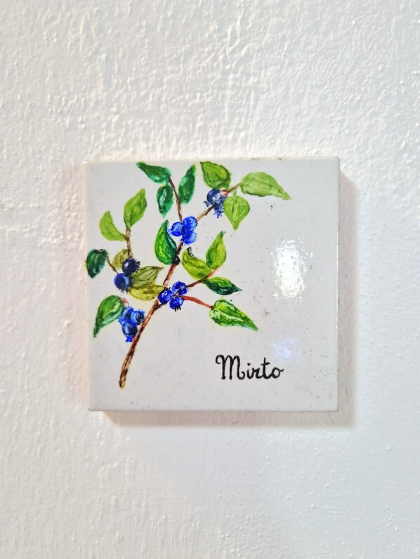 Un azulejo pequeño y cuadrado de cerámica con una rama de flores azules y hojas verdes, con la etiqueta 'Mirt'.