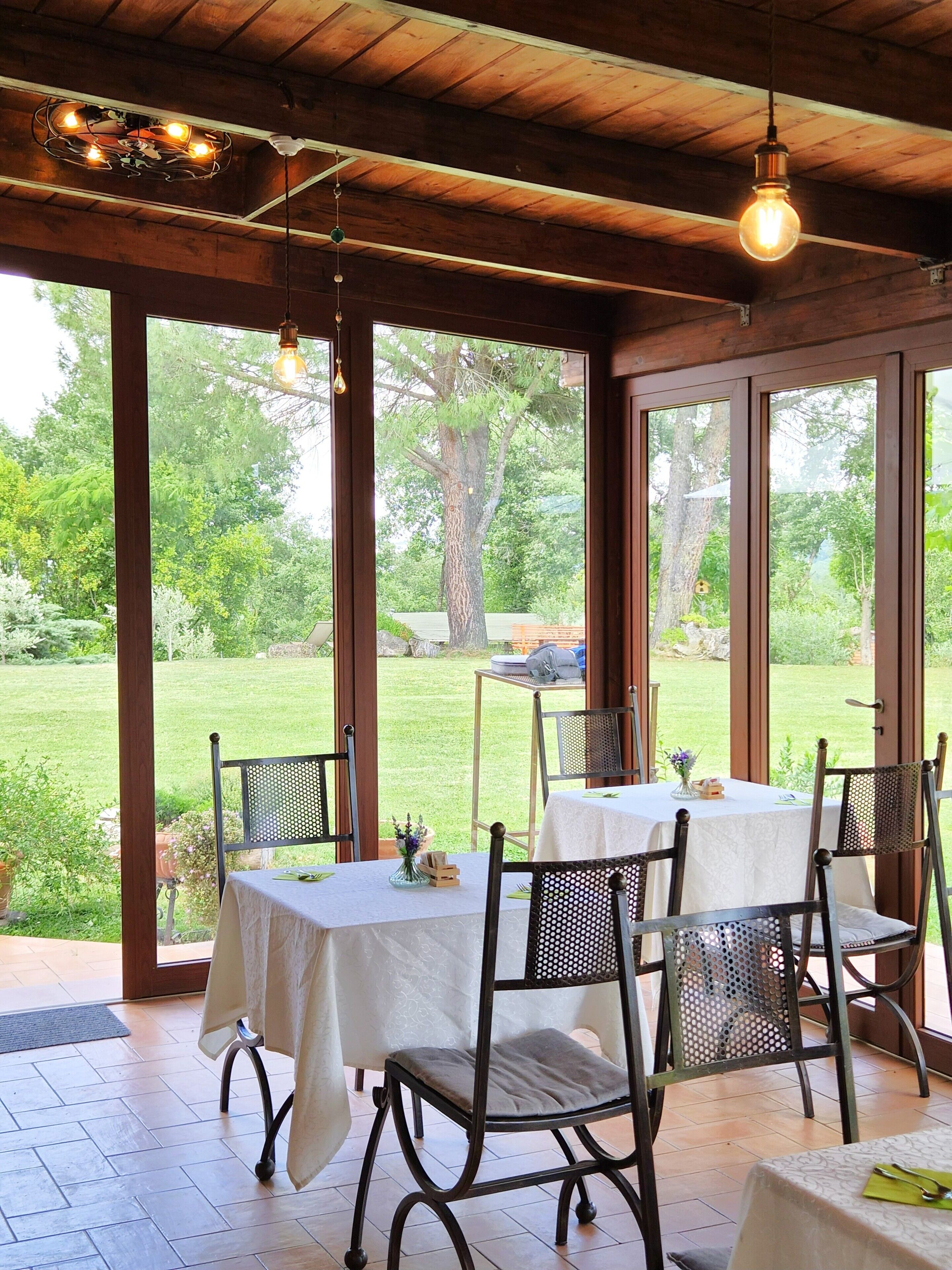 Un espacio de comedor al aire libre cubierto, con mesas y sillas, ventanas amplias y vistas a un paisaje verde.