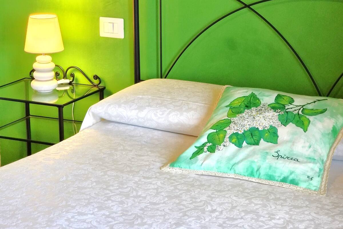 Una cama con una colcha blanca, una almohada verde con diseño floral, una mesita de noche con lámpara y una pared verde.