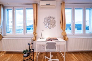 Ein Zimmer mit weißem Schreibtisch, einem Sessel, einem Heizkörper, einer Topfpflanze und einer Wand-Klimaanlage.
