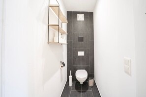 Une petite salle de bain aux carreaux sombres, avec des toilettes blanches, une poubelle et une étagère contenant des articles de toilette.