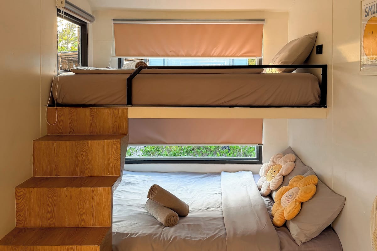 A bunk bed room with a window, wooden stairs, and a view of greenery outside.