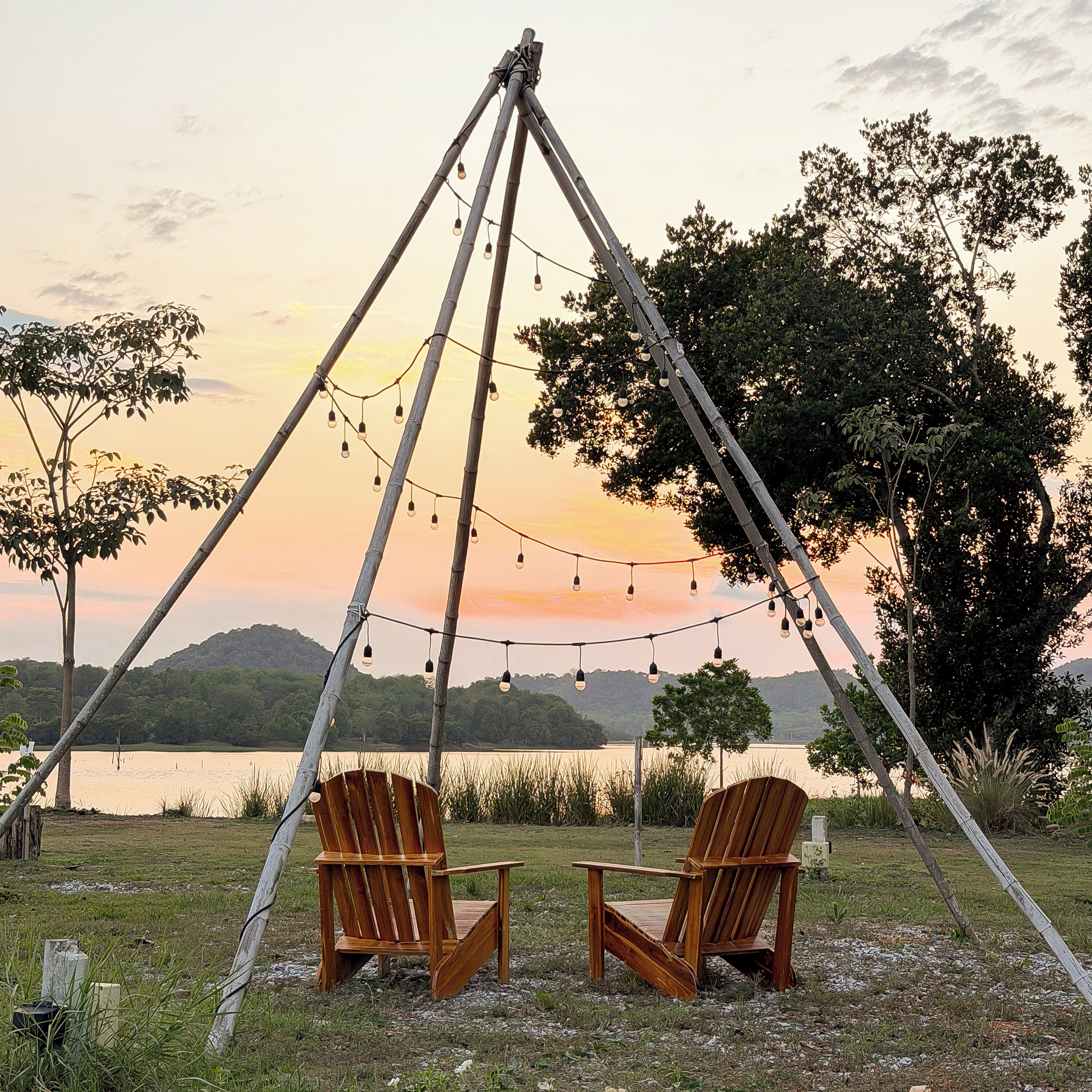 Dos sillas de madera bajo una estructura de bambú con luces colgantes, frente a una vista al atardecer de un lago y colinas.