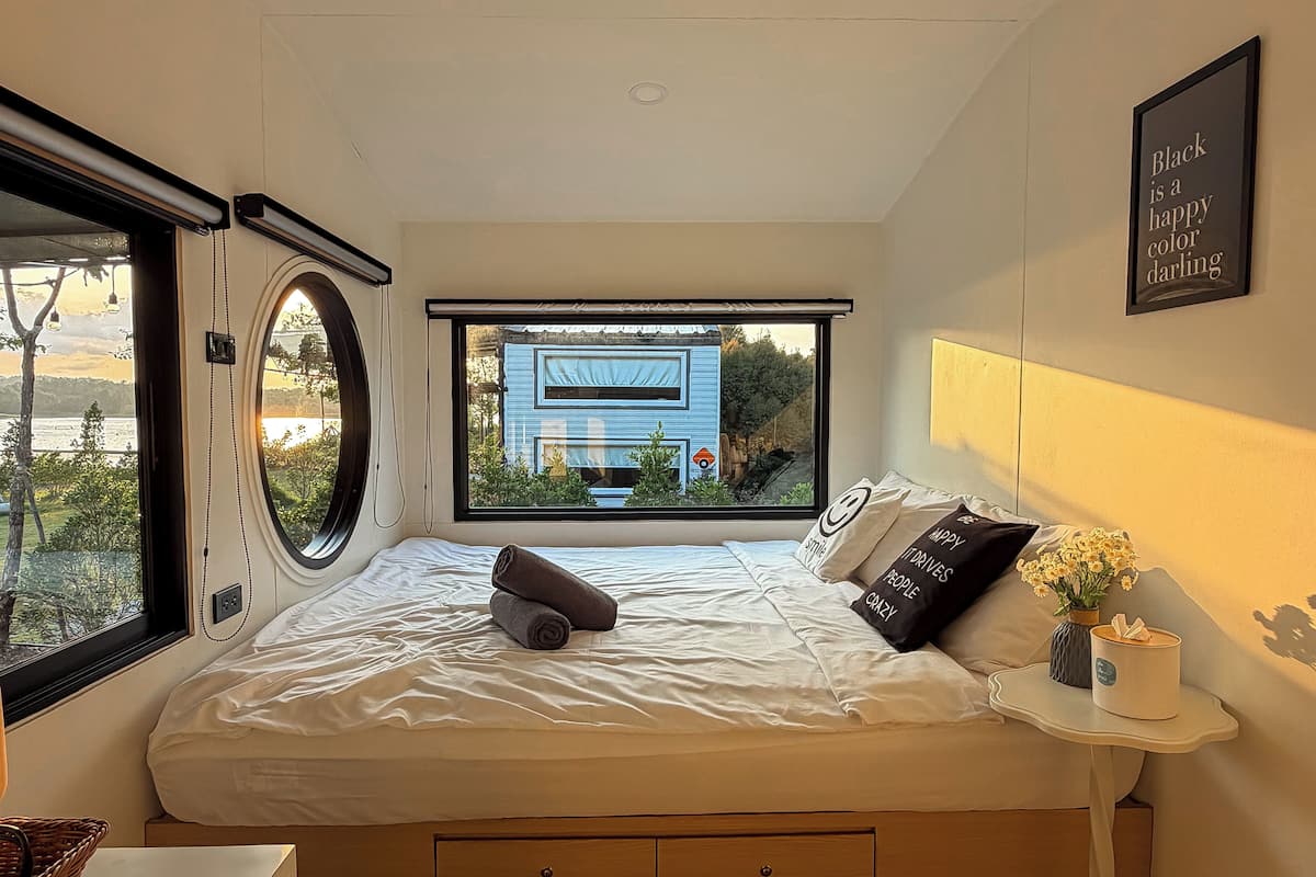 A bedroom with a large window, a bed with pillows, a bedside table with a vase, and a wall-mounted sign.