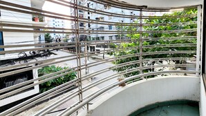 Un balcón curvo con barandilla de metal, vista a una calle con edificios y algo de vegetación.