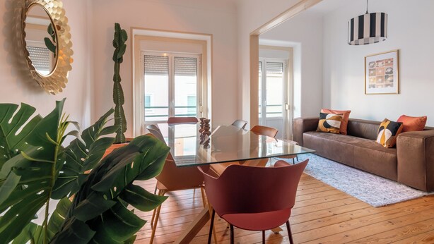 Ein modernes Wohnzimmer mit einem Glastisch, roten Stühlen, einem braunen Sofa und einer großen Grünpflanze.