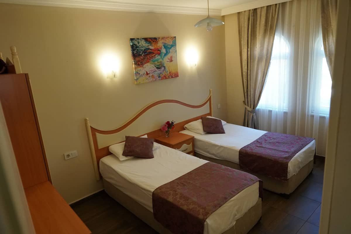 Ein Hotelzimmer mit zwei Einzelbetten, einem Holzkopfteil, einem Nachttisch mit einer Vase, einem Gemälde an der Wand und einem Fenster mit Vorhängen.