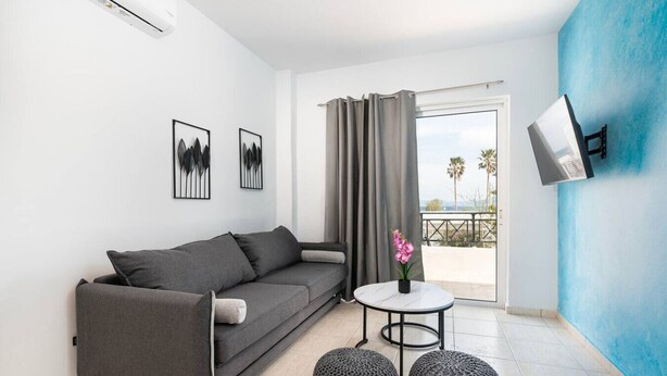 Een moderne woonkamer met een grijze bank, een klein rond tafeltje met een plant in pot en een televisie die aan een blauwe muur bevestigd is.