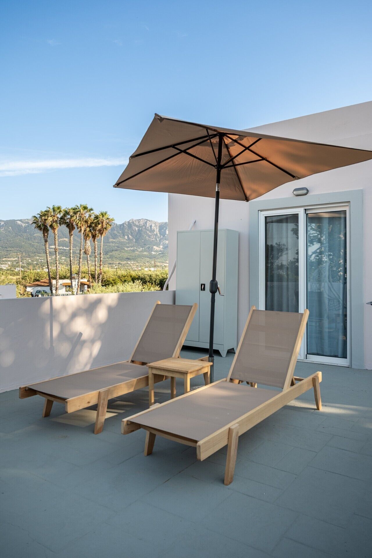A terrace with two wooden lounge chairs, a large umbrella, and a view of palm trees and mountains.