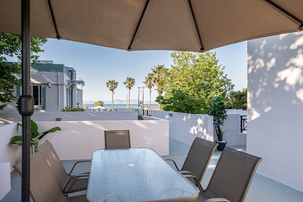 Eine Terrasse mit Tisch und Stühlen, einem großen Sonnenschirm und Blick auf das Meer und Palmen.