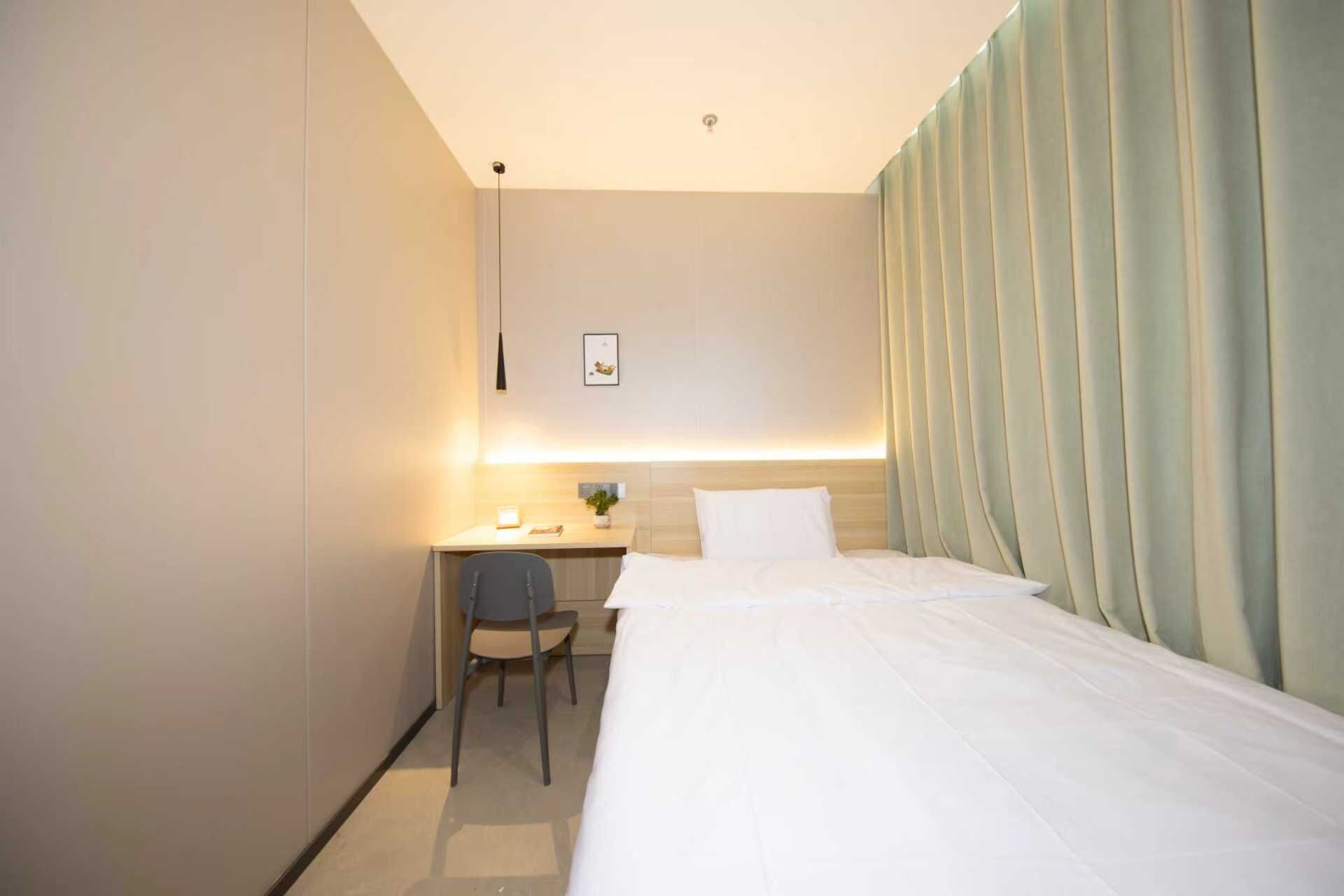 A hotel room with a bed, a desk, a chair, and a wall-mounted light fixture.