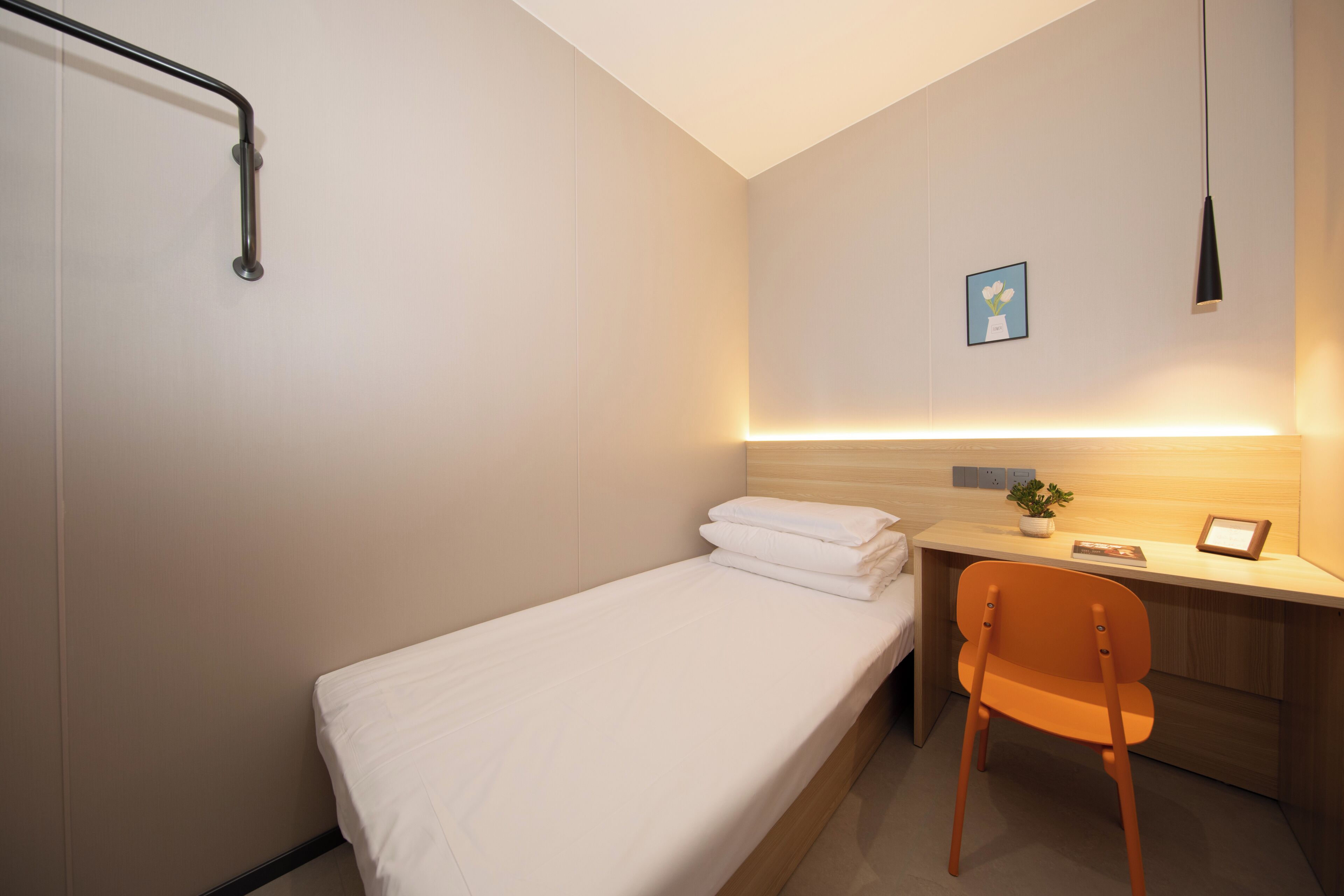 Ein Einzelbett mit weißer Bettwäsche, ein Holztisch mit Stuhl, ein kleiner Topfpflanze und ein gerahmtes Bild an der Wand.