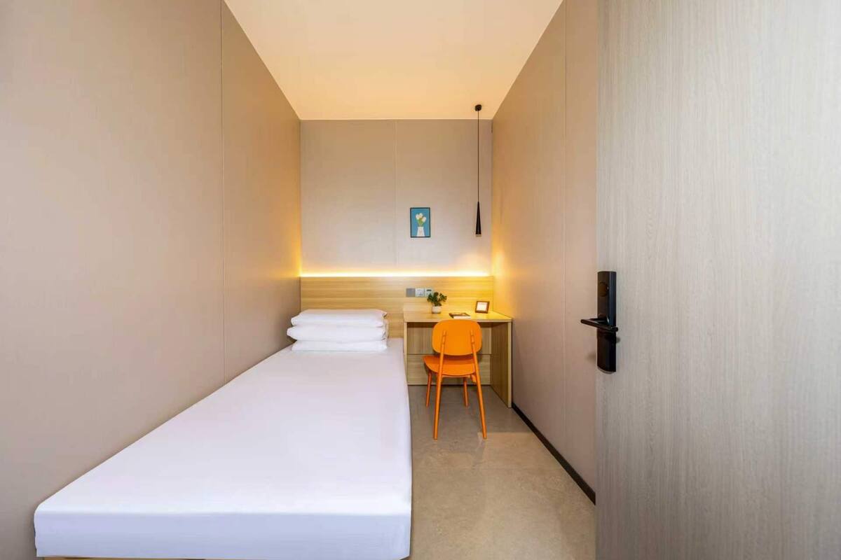 Una habitación pequeña con cama individual, escritorio y silla, mesita de noche y una lámpara fijada a la pared.