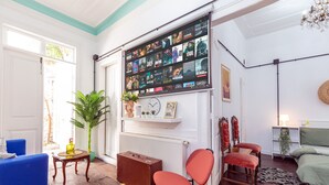 A living room with a large wall-mounted TV, a blue sofa, a red chair, a wooden table, and a suitcase.