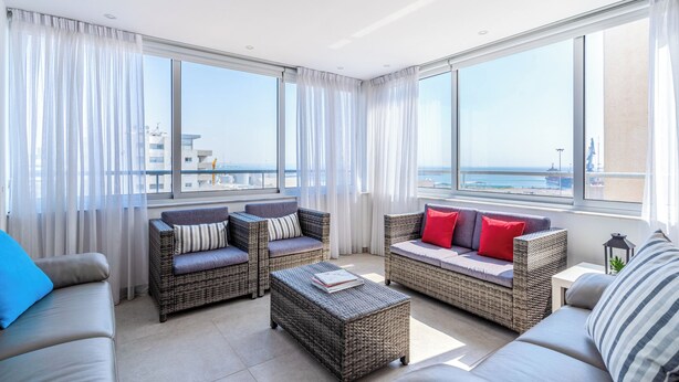 A spacious balcony with wicker furniture, large windows, and a view of the sea and buildings. - Panoramic 2-BR Apt & Balcony in Larnca (Larnaca)