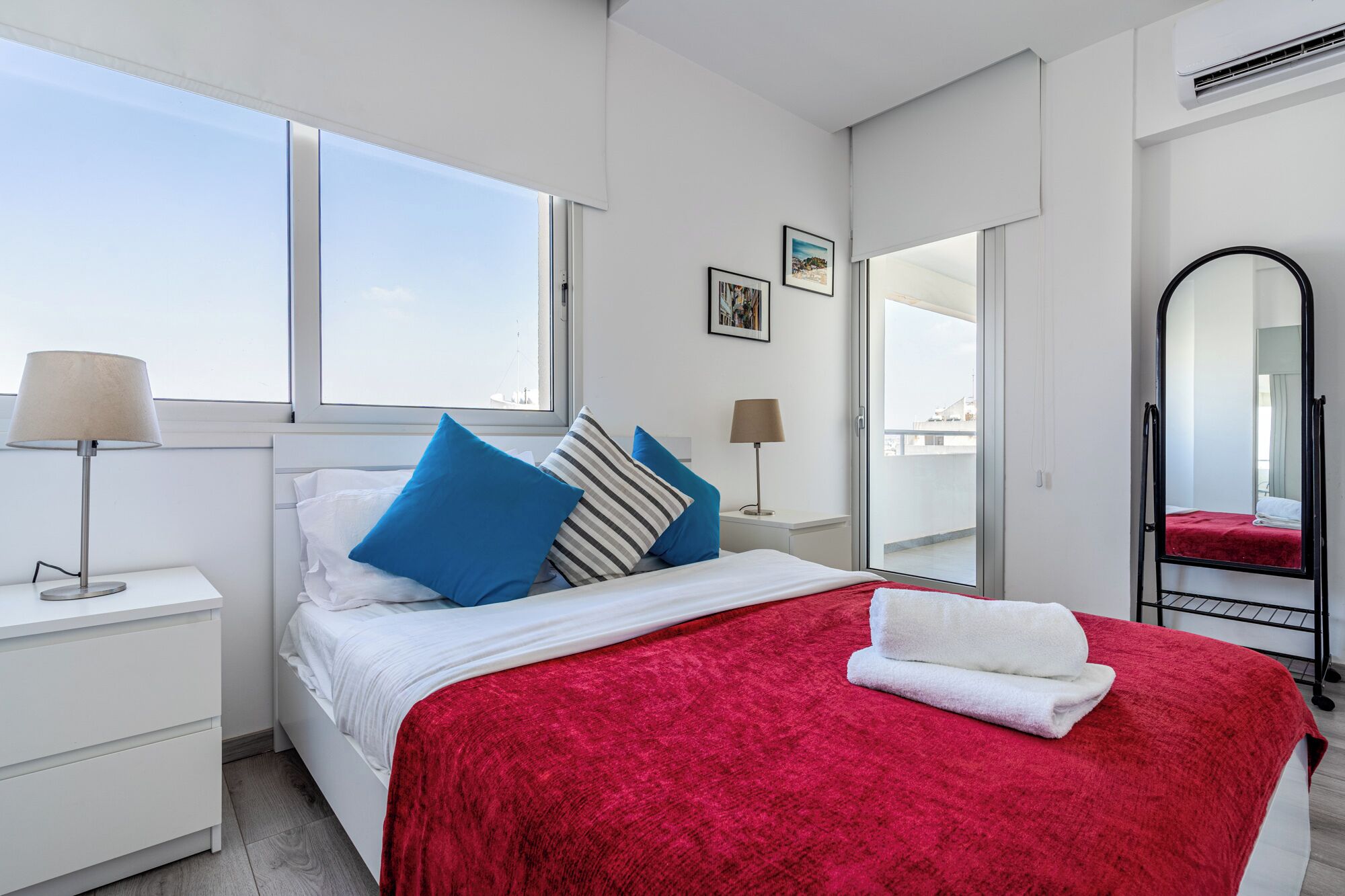 Une chambre à coucher avec un lit, une couverture rouge, une serviette blanche, une table de chevet, une lampe, une fenêtre et un miroir.