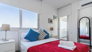 A bedroom with a bed, a red blanket, a white towel, a nightstand, a lamp, a window, and a mirror. - Panoramic 2-BR Apt & Balcony in Larnca (Larnaca)