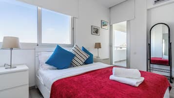 Quarto com cama, manta vermelha, toalha branca, mesa de cabeceira, abajur, janela e espelho.
